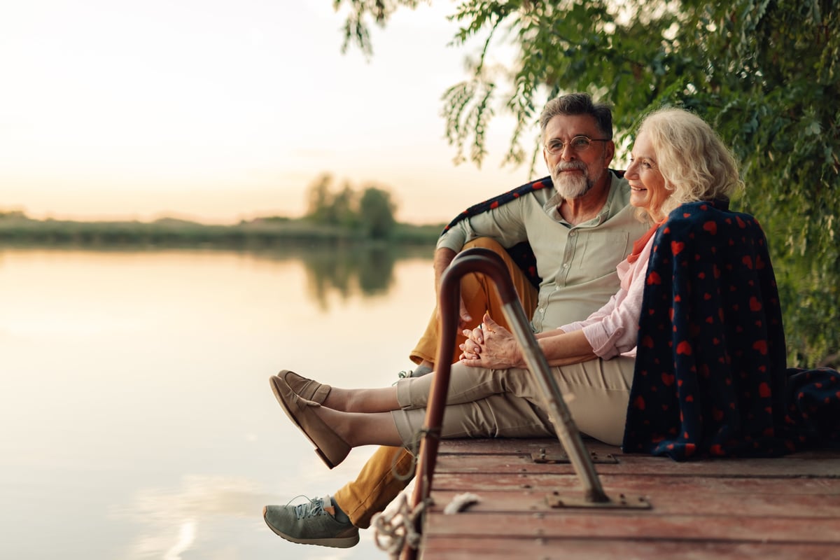 An older couple sitting together outdoors wrapped in a blanket, looking out calmly