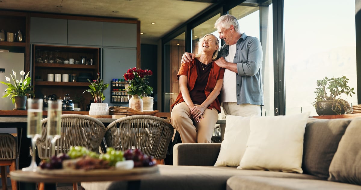 A couple in their sixties laughing together in the sunlit open kitchen of their own home, his arm around her shoulders
