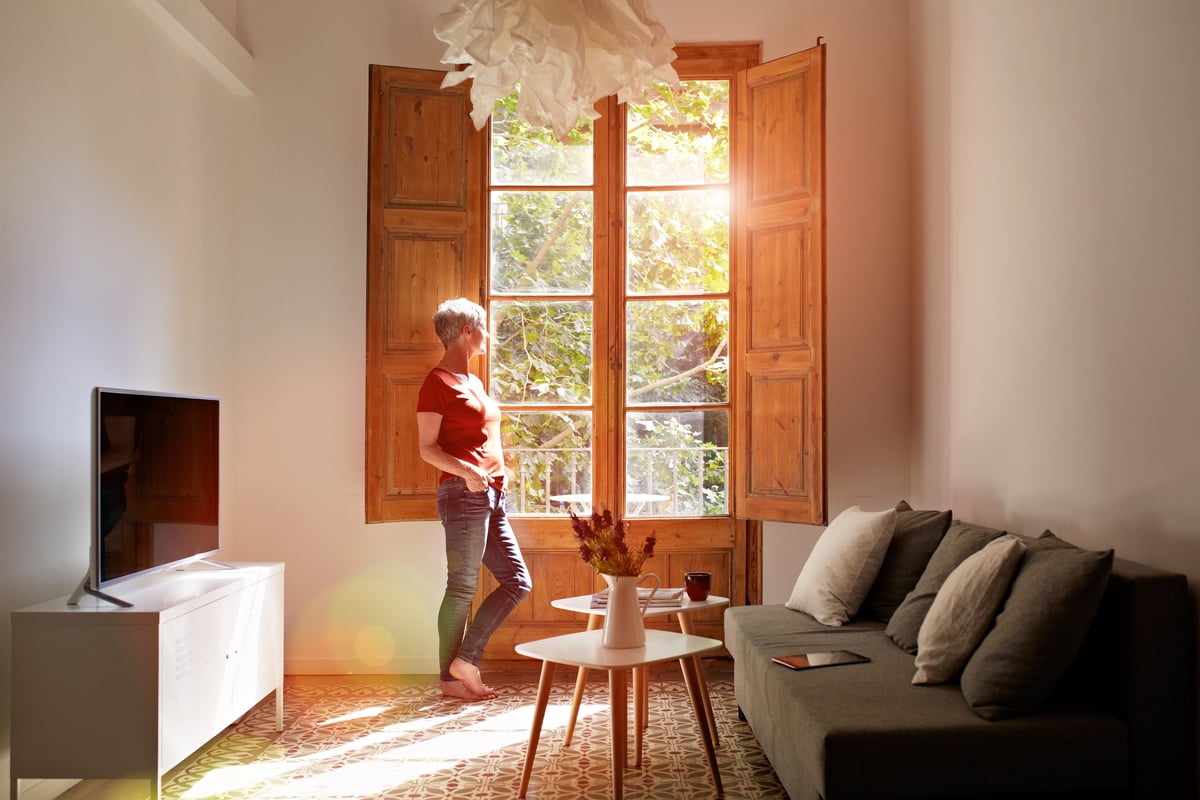 A woman in her seventies standing by a window in her home, looking out in thought
